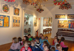 Dzieci siedzą na pufach w Sali wypełnionej ludowymi eksponatami
