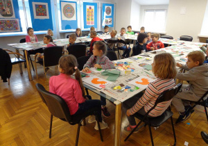 Dzieci siedzą przy stołach i wykonują symbol ludowy koguta