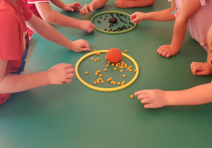 Dzieci siedzą przy stoliku. Na zdjęciu widać ich rączki. Segregują małe walce z darów zabawy do odpowiednich obręczy, w których znajdują się piłeczki z daru pierwszego oznaczające kolor zbioru