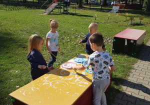 Czworo dzieci stoi przy skrzyni w ogrodzie przedszkolny. Na skrzyni położone są przybory do puszczanie baniek mydlanych. Dzieci maczają akcesoria w mydlinach