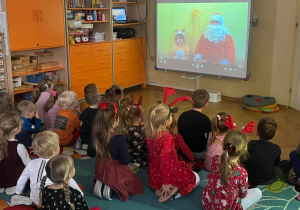 Dzieci siedzą na dywanie oglądając filmik wyświetlany na ekranie. Na ekranie widać Mikołaja i dziecko przebrane za renifera.