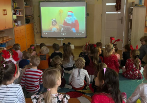 Dzieci siedzą na dywanie oglądając filmik wyświetlany na ekranie. Mikołaj na ekranie czyta dzieciom świąteczną bajkę.