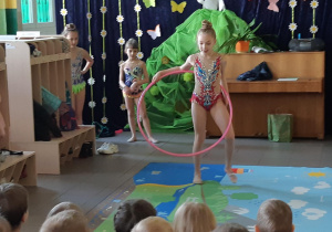Dziewczynka z hula-hop tańczy na dywanie. Dzieci oglądają jej występ.