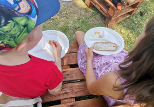 Dzieci siedzą na ławeczce w ogrodzie i spożywają śniadanie.