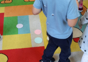 Dzieci maszerują po dywanie. Czekają na sygnał, aby wskoczyć na kolorowe kropki rozrzucone na dywanie.