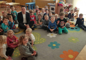 Dzieci siedzą na dywanie. Po środku, razem z dziećmi siedzi Przewodniczący Rady Miejskiej w Łodzi Pan Marcin Gołaszewski. Wszyscy uśmiechają się do zdjęcia.