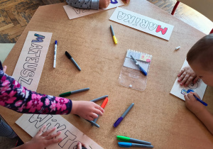 Dzieci pracuje przy stoliku, ozdabiają drewniane tabliczki ze swoim imieniem.