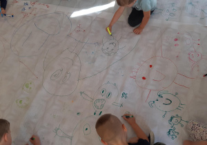 Na dywanie rozłożony duży arkusz papieru. Dzieci siedzą / leżą wokół i malują kolorowe koła, którym dorysowują twarze oraz nogi i ręce.