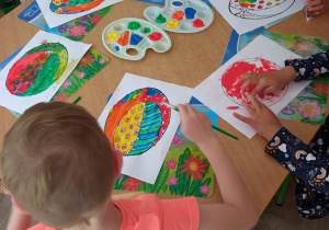 Dzieci siedzą przy stoliku i kolorują farbami rysunek piłki.