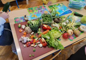 Na stoliku leżą różne warzywa. Dziewczynka przy stoliku obiera cukinię.