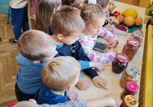 Dzieci stoją przy stoliku na którym leżą owoce oraz przetwory z owoców: dżemy, musy, kompoty.