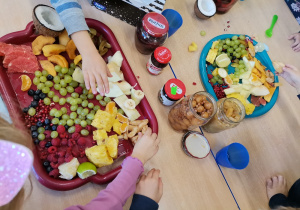 Na stoliku leżą tace z owocami oraz przetwory. Dzieci biorą do ręki owoce.