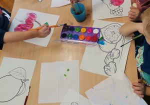 Dzieci siedzą przy stoliku - malują farbami owoce.