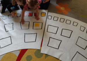 Dzieci na macie z narysowanymi kwadratami przekładają żółte i czerwone kartoniki.