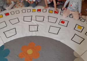 Dzieci siedzą wokół maty do kodowania. Na narysowanych kwadratach leżą rozłożone czerwone i żółte kartoniki.