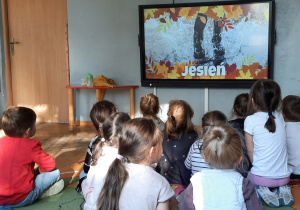 Dzieci siedzą na dywanie. Oglądają film edukacyjny o jesieni.