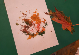 Dziecko odrywa z kartki pomalowanego farbami liścia. Na kartce widać odbitego liścia.