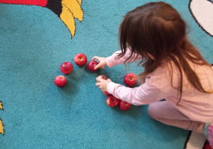 Dziewczynka na dywanie układa z jabłek cyfrę 2.