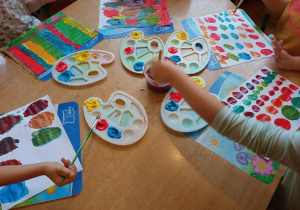 Dzieci siedzą przy stoliku, malują farbami, mieszając ze sobą różne kolory.