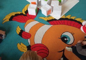Dzieci siedzą w kole na dywanie, na środku koła leżą kwadratowe pudełka z pomarańczowymi i zielonymi plakietkami.