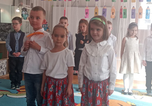 Dzieci na dywanie stoją do występu. Dziewczynki w białych bluzkach i czerwonych spódniczkach w kwiatuszki.