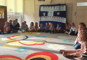 Dzieci podczas pasowania siedzą w półkolu na tle dekoracji Pasowanie na Przedszkolaka.