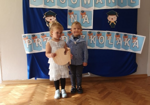 Chłopiec i dziewczynka stoją na tle dekoracji Pasowanie na Przedszkolaka. Dziewczynka trzyma dyplom.