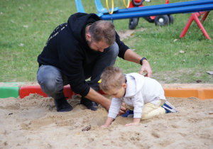 Chłopiec bawi się w piaskownicy. Obok niego kuca mężczyzna.