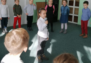 Dziewczynka stoi na środku sali z mikrofonem. Pozostałe dzieci elegancko stoją w sali.