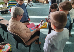 Dzieci wręczają kartki z życzeniami siedzącej na krześle mieszkance DPS – u.
