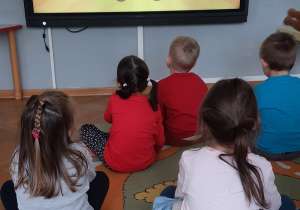 Dzieci siedzą na dywanie i oglądają film o Dniu Pluszowego Misia