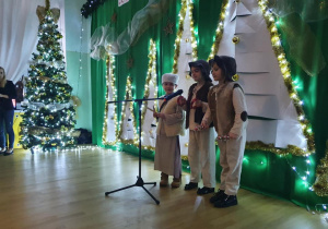 Troje dzieci występuje na scenie. W tle dekoracja. Białe choinki na zielonym tle.