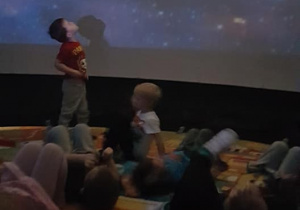 Dzieci w środku planetarium oglądają uśmiechnięte, animowane planety. Jeden z chłopców stoi, aby przyjrzeć się bliżej.