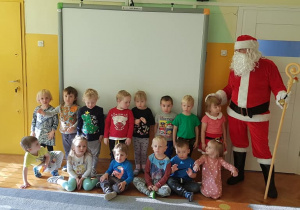 Dzieci z grupy żółtej są na scenie z Mikołajem. Obserwują Mikołaja, oglądają czekoladowe Mikołaje, uśmiechają się, machają rączkami.