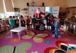 Dzieci z grupy fioletowej pozują do zdjęcia z Mikołajem. Machają do publiczności i uśmiechają się ze sceny, Towarzyszy im nauczycielka.
