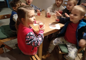 Dzieci siedzą przy stoliku, piją truskawkowy soczek ze szklanek.