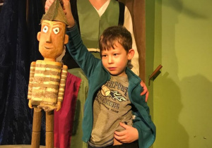 Chłopiec stoi na scenie i trzyma dużą marionetkę, pomaga mu w tym aktorka.