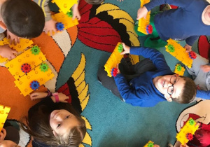 Dzieci układają klocki na dywanie, niektóra z nich siedzą, inne leżą.