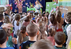 Dzieci śpiewają piosenkę i powtarzają ruchy za kobietą stojącą na scenie.
