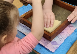 Dziewczynka gąbką odsącza wodę, aby powstał papier czerpany.