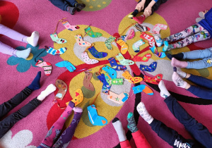Na środku dywanu ułożone są kolorowe, papierowe skarpetki. Dookoła nich siedzą dzieci pokazują na swoich stopach skarpetki nie od pary.