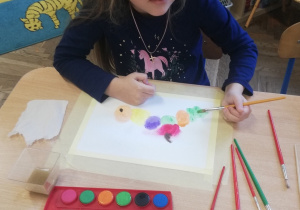 Dziewczynka maluje kompozycje owoców farbami akwarelowymi.