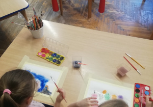 Dwie dziewczynki malują farbami owoce na papierze akwarelowym.