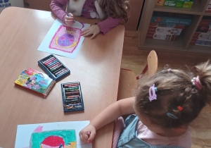 Dwie dziewczynki siedząc przy stole rysują pastelami olejnymi na białej kartce.