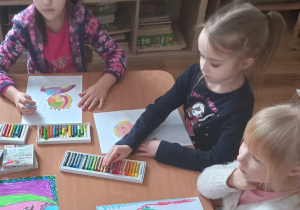 Trzy dziewczynki rysują portrety pastelami na białych kartkach.