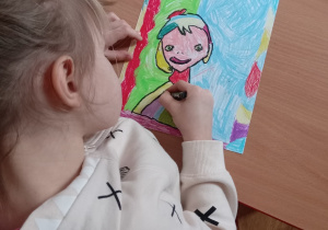 Dziewczynka rysuje kontury czarnym pastelem na wykonanym portrecie.