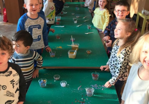 Stoły w sali są połączone w jeden długi. Po oby stronach stoją dzieci, uśmiechają się do zdjęcia. Na stole stoją szklaneczki do wyrobu świec.