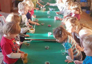 Dzieci stoją po obu stronach stołu. Za pomocą łyżeczek nabierają ozdoby i przesypują do szklaneczek.