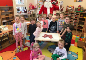 Dzieci z grupy czerwonej stoją na dywanie. Za nimi stoi Mikołaj. Między dziećmi znajduje się stolik – prezent od Mikołaja. Na dywanie siedzą chłopiec i dziewczynka.