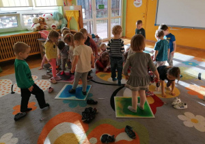 Dzieci bawią się prezentem od Mikołaja. Na dywanie leżą kolorowe kafle sensoryczne. Dzieci zdejmują buty. Wchodzą na kafle stopami.